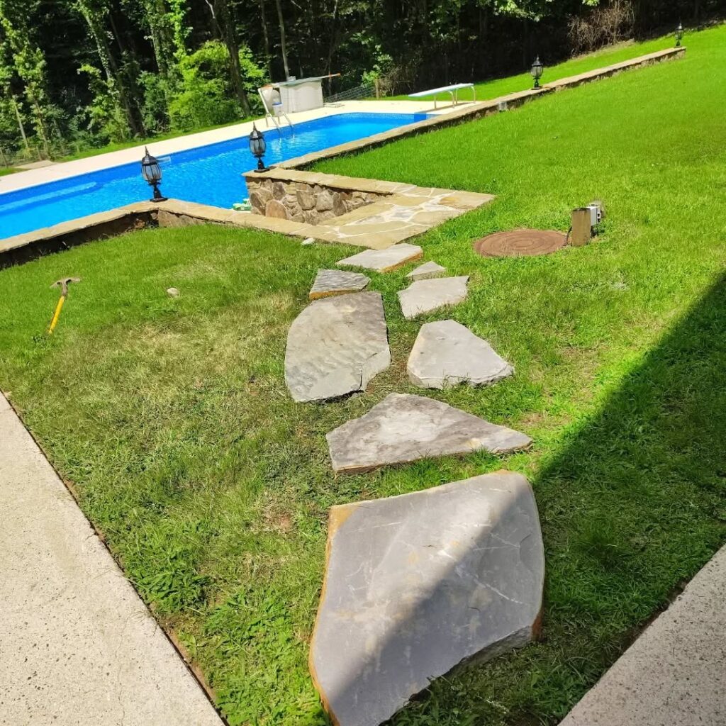 Large flat stones form a path across grass leading to a swimming pool bordered by stone and green lawn.